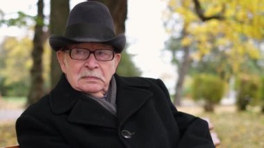 Siyah ceketli gri bıyıklı düşünceli bir son sınıf öğrencisi parkta bir bankta oturuyor.