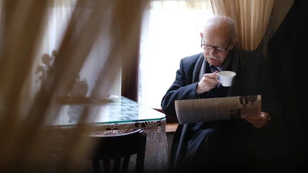Homem sênior segurando uma xícara de café sentado em uma mesa em um café lendo um jornal. — Fotografia de Stock