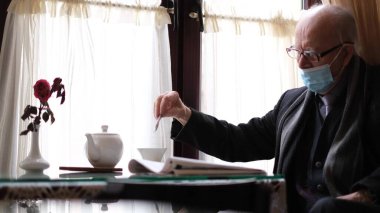 Koruyucu maskeli yaşlı bir emekli kafede otururken çayı karıştırıyor.