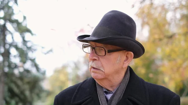 Un viejo abuelo en gafas y un sombrero negro camina en el parque de otoño de la ciudad —  Fotos de Stock