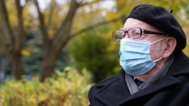 Tıbbi koruyucu maskeli ve gözlüklü kıdemli bir adam parkta dinleniyor..