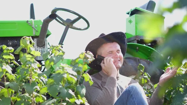 Un joven agricultor usa un teléfono inteligente, está descansando cerca de su tractor. —  Fotos de Stock