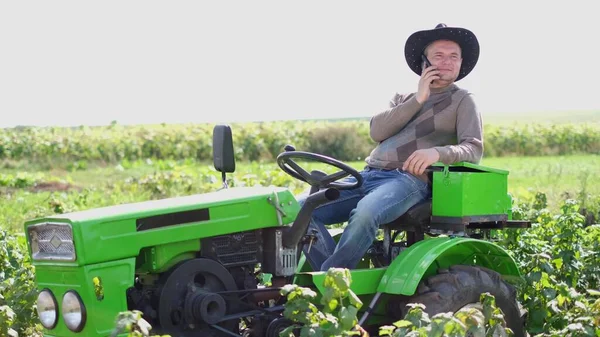 Giovane agricoltore seduto su un trattore verde. L'uomo del villaggio che parla al telefono. — Foto Stock