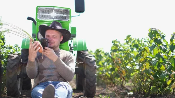 Jordbrukare inspekterar fotografera morot skörd på smartphone medan du sitter nära sin traktor — Stockfoto