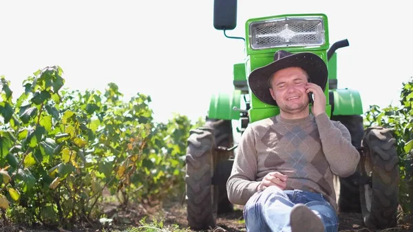 Um jovem agricultor usa um smartphone, ele está descansando perto de seu trator. — Fotografia de Stock