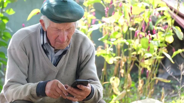 Grand-père aux cheveux gris utilise un smartphone assis sur une chaise près de la maison. — Photo