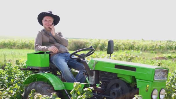 Young farmer sitting on a tractor and talking on a smartphone in the field. — Stock Video