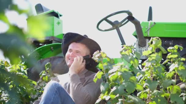 Un joven agricultor se comunica en un teléfono inteligente cerca de su tractor. — Vídeo de stock