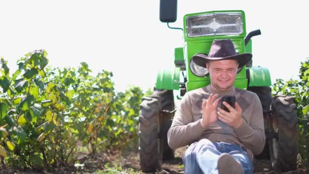 Um jovem agricultor se comunica em um smartphone perto de seu trator. — Vídeo de Stock