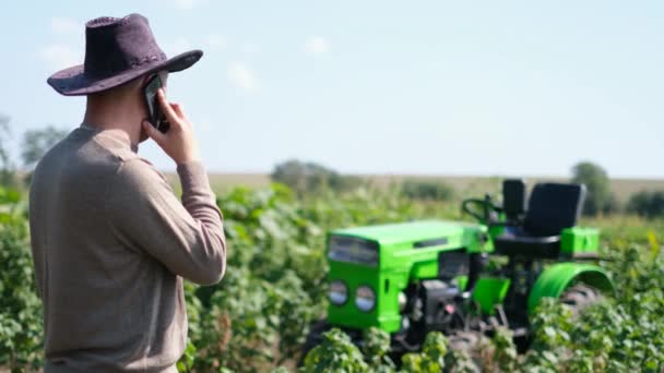 Un granjero usa un teléfono inteligente en un campo cerca de su tractor. — Vídeo de stock