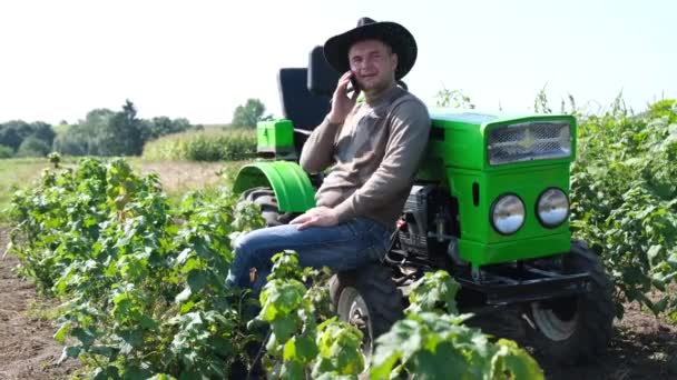 Biały farmer rozmawiający przez telefon stojący przy zielonym traktorze. Działalność rolnicza. — Wideo stockowe