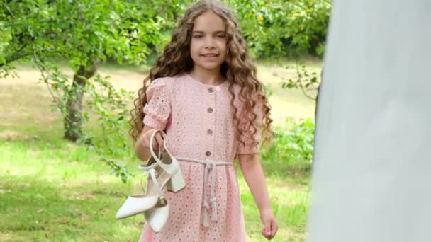 Το κοριτσάκι με τις μπούκλες φοράει παπούτσια στις νύφες. Παιδί στον κήπο κοντά στο σπίτι. — Αρχείο Βίντεο