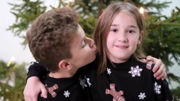 Младший брат целует сестру в щеку. Счастливая семья на фоне елки — стоковое видео