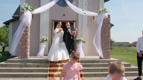 A beautiful young couple comes out of the church and scatters candy to the children. — Stock Video