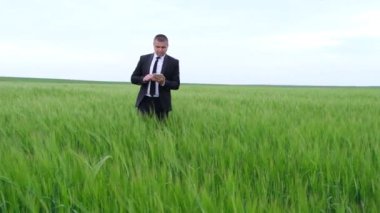 Takım elbiseli genç işadamı elinde tabletle buğday tarlasında yürüyor.,