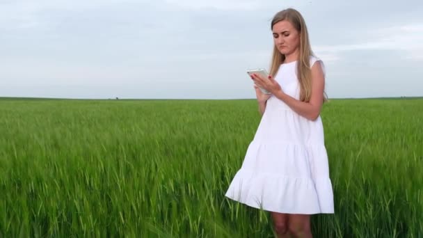 Ung flicka i en vit klänning med en tablett i händerna står i ett grönt fält av vete. — Stockvideo