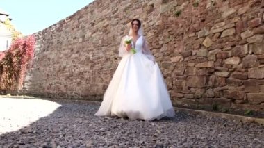 Lüks elbiseli gelin, taştan bir duvarın arkasında yürüyor ve gülüyor.