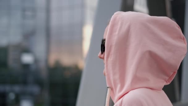 Una chica con una capucha en la cabeza mira el atardecer. Mujer de moda en una chaqueta rosa. — Vídeos de Stock