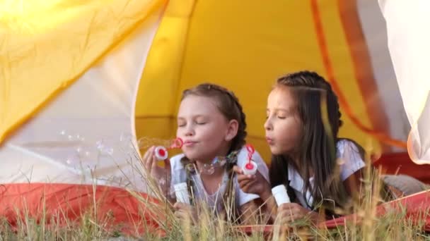 Dos niñas son hermanas, están sentadas en una tienda de campaña y soplando burbujas. — Vídeos de Stock
