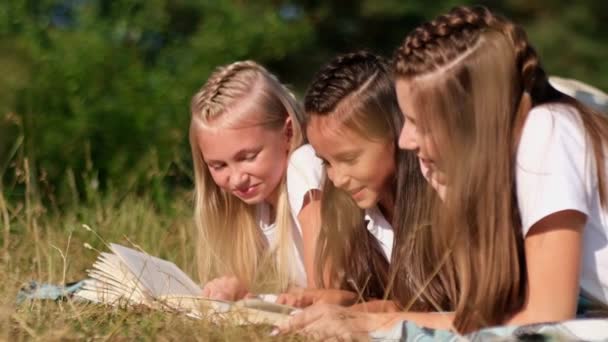 Les adolescentes lisent un livre allongé sur l'herbe verte, elles s'amusent dans un camp d'été. — Video