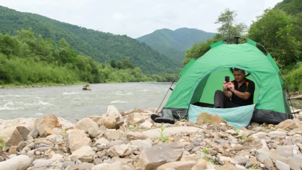 산에서 강을 따라 쉬고 있는 남자가 녹색 텐트에 앉아 스마트폰을 사용하고 있습니다. — 비디오