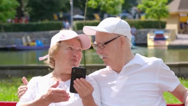Pár v důchodu sedí na lavičce v parku a učí se nový smartphone. — Stock video