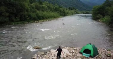 Bir balıkçı dönerek balık tutar, dağ nehri manzarası ve yeşil bir çadır.