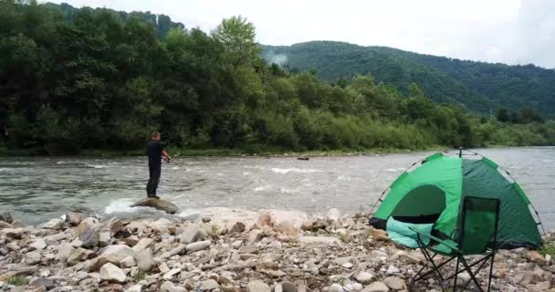 Di tepi sungai pegunungan, seorang nelayan menangkap ikan, memasang tenda hijau. — Stok Video