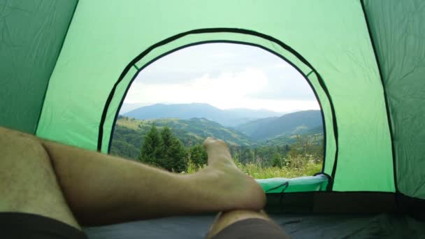 Парень-турист отдыхает, лежа в палатке, красивый вид из палатки на горы, летнее время — стоковое видео