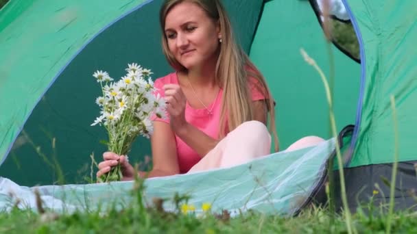 Hermosa chica rubia sentada en una tienda de campaña, ella sostiene flores rotas. Senderismo en las montañas — Vídeos de Stock