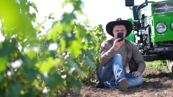 En ung bonde snakker i telefonen på en vingård med en traktor i nærheten.. – stockvideo