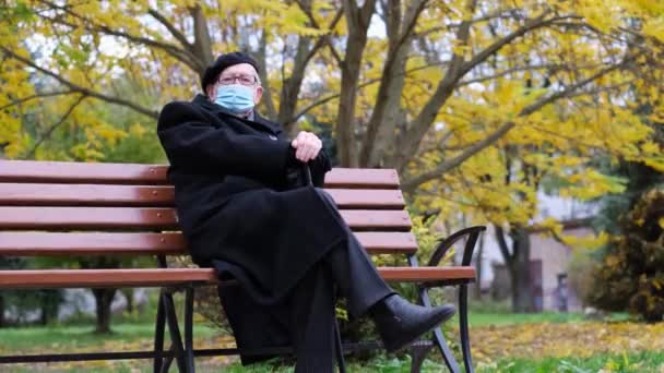Egy öreg nagyapa fekete kabátban és svájcisapkában, egy padon ül egy védőmaszkban. — Stock videók