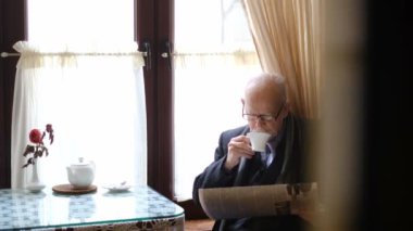 Gri bıyıklı emekli son sınıf öğrencisi mutfakta gazete okuyor.