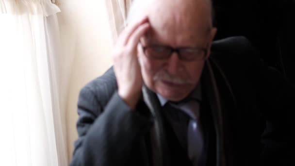 Een oudere grijze man die op een stoel zit in een kamer bij het raam, zijn hoofd doet pijn. — Stockvideo