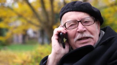 Yaşlı bir dede akıllı bir telefonla konuşuyor, parkta bir bankta oturuyor.. 