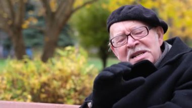 Yaşlı adam kameraya konuşuyor, ellerini göstermesini açıklıyor. Emekli adam parkta oturuyor.