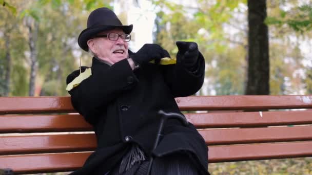Un anciano está sentado en un banco del parque, mirando hojas amarillas. Vida de jubilación — Vídeos de Stock
