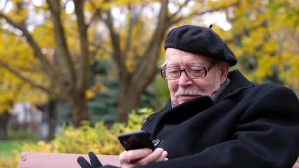 Starý děda mluví na smartphonu, sedí na lavičce v parku. — Stock video