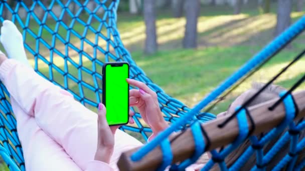 Mujer descansando acostada en una hamaca en el bosque, ella sostiene un teléfono inteligente en sus manos con un cromakey — Vídeos de Stock