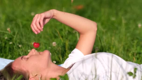 Το κορίτσι με το άσπρο μπλουζάκι ξεκουράζεται, ξαπλωμένο στο πράσινο γρασίδι, τρώει κόκκινα ζουμερά κεράσια. — Αρχείο Βίντεο