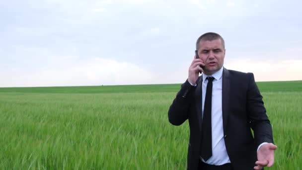 Hombre de negocios enojado hablando en un teléfono inteligente en medio del campo. Hombre con traje negro — Vídeos de Stock