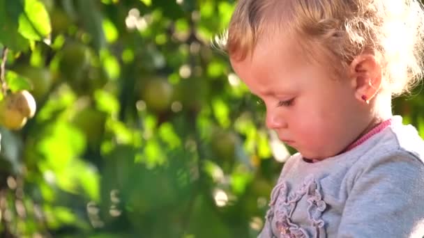 Egy szőke hajú kislány pihen a kertben, egy gyermek a szabadban egy almafa mellett.. — Stock videók