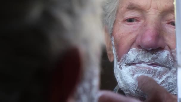 El abuelo se afeita cerca del espejo, se unta la cara con espuma blanca. — Vídeo de stock