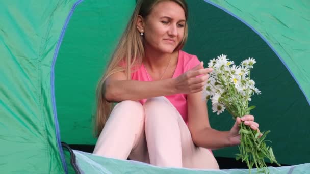 Дівчина відпочиває в наметі в горах, вона тримає квіти ромашки в руках . — стокове відео
