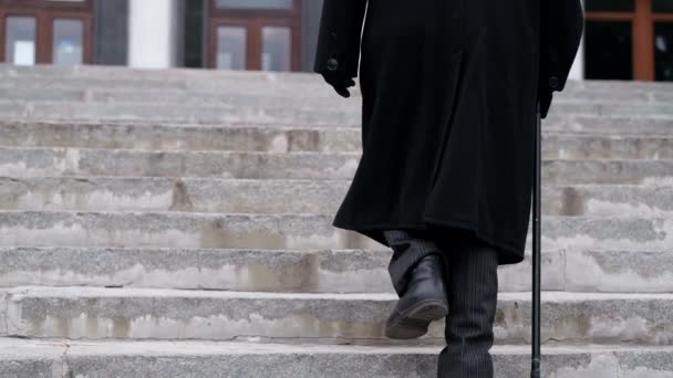 Старый дед в черном пальто поднимается по лестнице, идя тяжело. 80-летний пенсионер — стоковое видео