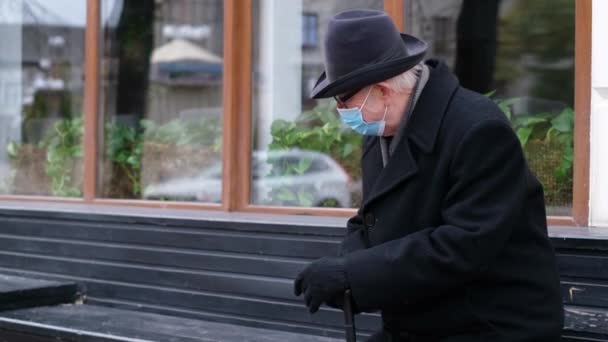 El pensionista descansa en un banco de la ciudad durante la cuarentena. Abuelo en una máscara protectora — Vídeos de Stock