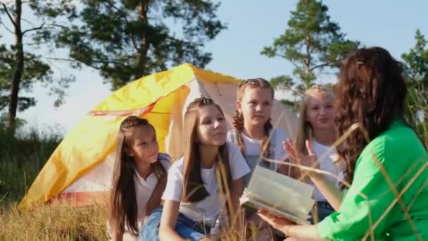 Un joven profesor lee un libro a las niñas en un campamento de verano a través del trabajo de un educador. — Vídeos de Stock