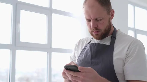 Der junge Künstler in einem weißen Atelier blickt auf sein Smartphone. Ein Künstler in grauer Schürze bereitet sich vor — Stockfoto