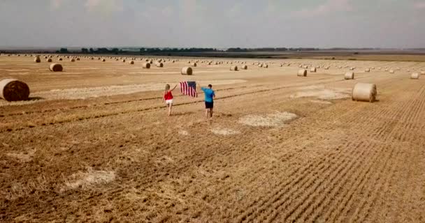 Un jeune couple heureux court insouciant et saute sur un champ de blé. Tenant le drapeau des États-Unis — Video