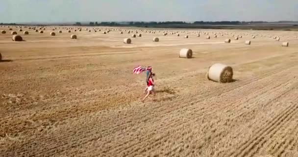 Młoda szczęśliwa para biegnie beztrosko i skacze na polu pszenicy. Posiadanie flagi USA — Wideo stockowe